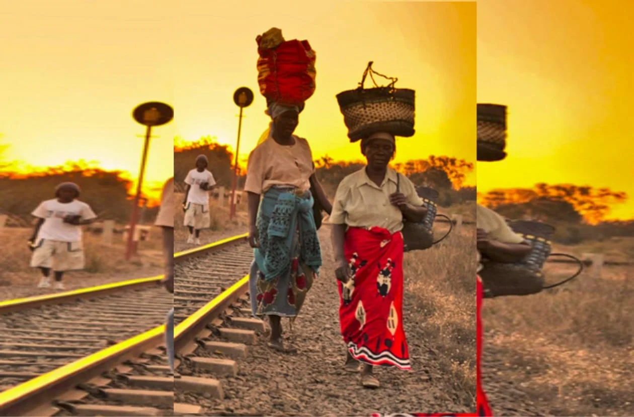 Women in colourful cloth wraps carrying baskets on their heads along a railway line at sunset